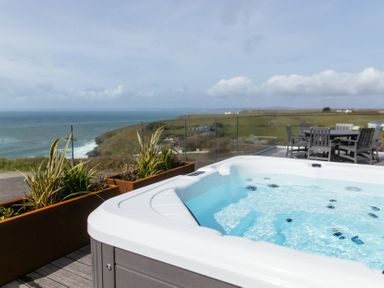 A hot tub on a deck with potted plants and outdoor dining furniture overlooking the ocean and coastline at Shearwater in Mullion
