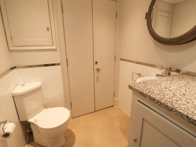 A bathroom with a toilet, sink, round mirror, cabinet, and tiled floor at Sea Crest in Aldeburgh