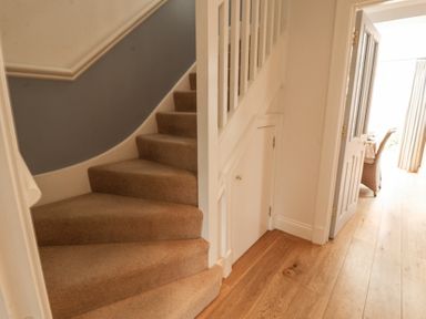 A carpeted staircase next to a small door under the stairs and a hallway with wooden floor and open door at Sea Crest in Aldeburgh
