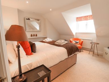 A bedroom with a double bed orange and brown cushions a lamp a small side table an orange armchair a window with orange patterned blinds and a wall mirror at Sea Crest in Aldeburgh