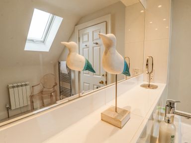 A bathroom countertop with a bird sculpture soap dispenser and small mirror in front of a large mirror at Sea Crest in Aldeburgh