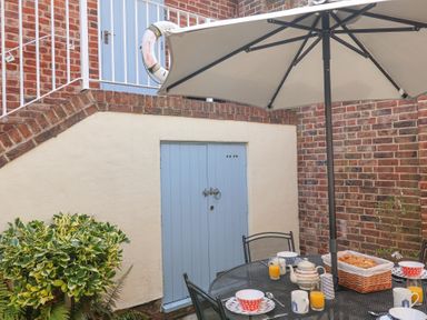 An outdoor patio with a black metal table and chairs under a large umbrella with breakfast items near a blue door at Sea Crest in Aldeburgh