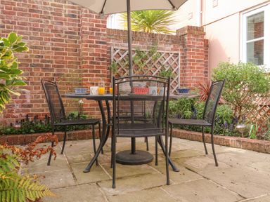 An outdoor patio area with a metal table and four chairs under an umbrella surrounded by brick walls and plants at Sea Crest in Aldeburgh