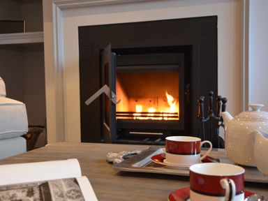 A fireplace with fire burning and a coffee table holding a white teapot and two red and white cups at Sea Crest in Aldeburgh