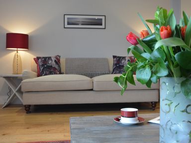 A beige sofa with patterned cushions and a red lamp on a side table in a living room with a coffee table and flowers in a vase at Sea Crest in Aldeburgh