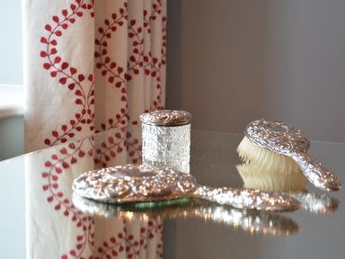 A decorative silver hairbrush set with a jar on a reflective surface near a curtain with red leaf patterns at Sea Crest in Aldeburgh