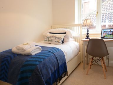 A bedroom with a single bed a blue blanket folded towels a desk with a laptop and a lamp and a chair at Sea Crest in Aldeburgh