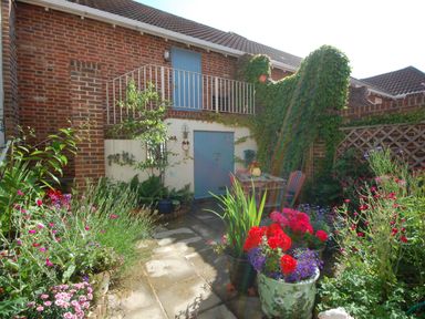 A small garden with various flowering plants pots patio furniture and a brick building with blue doors and ivy at Sea Crest in Aldeburgh