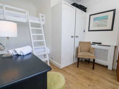 A small bedroom with white bunk beds a black desk with a lamp and towels a beige chair and a white wardrobe at Flat 7 Sand Acres in Canford Cliffs
