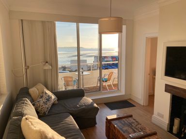 A living room with a gray sofa pillows wooden chest and television near a balcony overlooking the sea at Flat 7 Sand Acres in Canford Cliffs