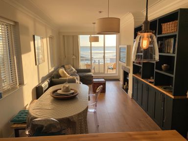 A dining area with a table and chairs next to a living room with a sofa and a bookshelf overlooking a balcony with a sea view at Flat 7 Sand Acres in Canford Cliffs