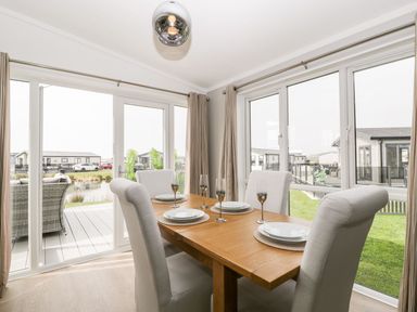 A dining area with a wooden table set with four plates and glasses surrounded by upholstered chairs with large windows overlooking a garden and neighboring houses at Summerdown Brean