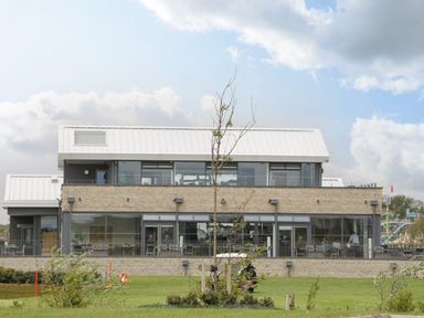 A modern two-story building with large glass windows and a metal roof surrounded by grass and small trees at Summerdown in Brean