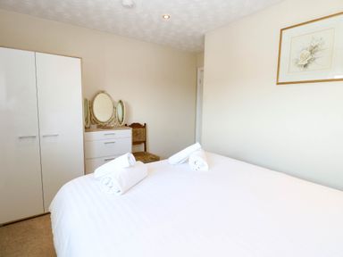 A bedroom with a white bed and rolled towels a wardrobe a dresser with a tri-fold mirror and a chair at Sweetacres in Hemsby