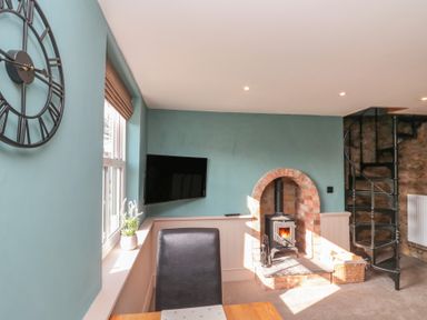 A room with a wall clock window with plant wall mounted television wood burning stove and spiral staircase at Top Holme Cottage in Newbald