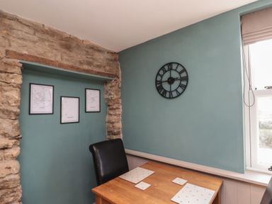 A dining area with a wooden table set with placemats a black chair a stone wall with framed maps on a teal wall and a window with a blind at Top Holme Cottage in Newbald