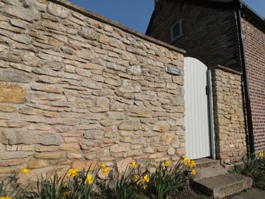 A stone wall with yellow flowers in front and a white wooden gate at Top Holme Cottage in Newbald