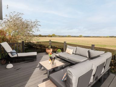 An outdoor patio with grey cushioned sofas, lounge chair, small table with flowers and bottle overlooking an open field at The Gallops in Chipping Norton