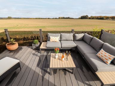 An outdoor deck with a grey corner sofa, a wooden coffee table with flowers and drinks, and potted plants at The Gallops in Chipping Norton
