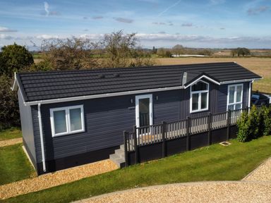 A single story dark grey house with white trim and a small fenced porch surrounded by grass and gravel paths at The Gallops in Chipping Norton