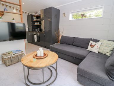 A living room with a gray sectional sofa and a wooden round coffee table with candles in front of a kitchen with dark cabinets and a wall-mounted TV at The Gallops in Chipping Norton