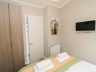 A bedroom with a bed with folded towels a wooden wardrobe a white door a wall-mounted TV and a painting of a jockey on a horse at The Gallops in Chipping Norton
