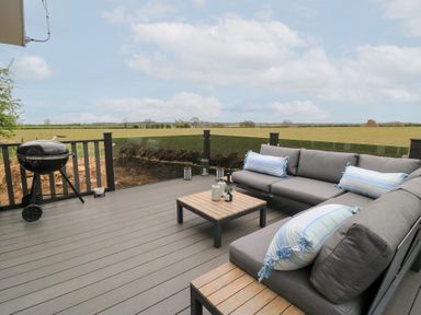 An outdoor deck with a gray sectional sofa with pillows a wooden coffee table and a charcoal grill overlooking a field at The Gallops in Chipping Norton