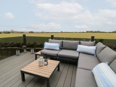 An outdoor seating area with a grey sectional sofa and wooden coffee table on a deck overlooking a field at The Gallops in Chipping Norton