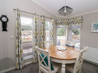 A dining area with a round wooden table four chairs with cushions patterned curtains and blinds on windows at Lodge 2 in Auchterarder
