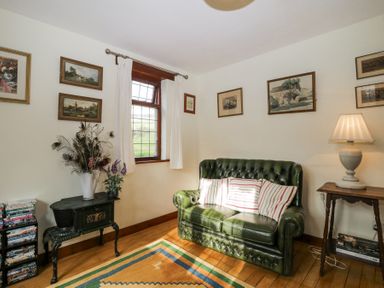 A small living room with a green leather sofa striped pillows a lamp on a wooden side table framed paintings on white walls and a decorative stove at Wringford Farm Annexe Rame near Kingsand