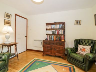 A room with a wooden bookshelf filled with books a wooden side table with a white lamp two green leather armchairs and a colorful rug on the floor at Wringford Farm Annexe Rame near Kingsand