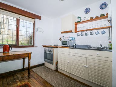 A kitchen with a wooden table and kettle by a window a stove and oven a microwave sink and shelves with cups and glasses at Wringford Farm Annexe in Rame near Kingsand