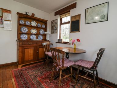 A dining area with a round wooden table with three wooden chairs with cushions and a wooden cabinet with blue and white plates at Wringford Farm Annexe in Rame near Kingsand