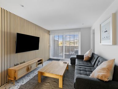 A living room with a dark gray sofa two pillows a wooden coffee table a wooden TV stand with a mounted flat screen TV and a large window with blinds at 5 Mermaid Court in Newquay
