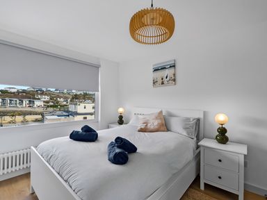 A bedroom with a bed with white bedding and blue towels on it two bedside tables with lamps and a window with a view of buildings at 5 Mermaid Court in Newquay