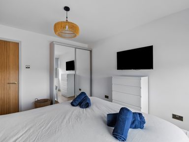 A bedroom with a bed with blue towels a white dresser a wall-mounted TV a closet with mirrored sliding doors a wooden door and a wicker basket at 5 Mermaid Court in Newquay