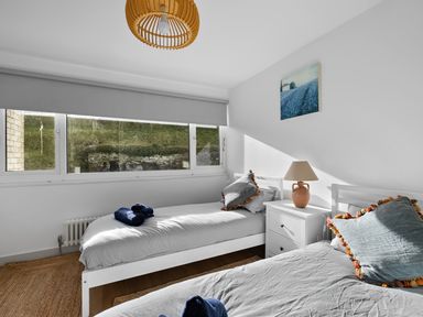 A bedroom with two single beds side by side a white nightstand with a lamp and a painting on the wall at 5 Mermaid Court in Newquay