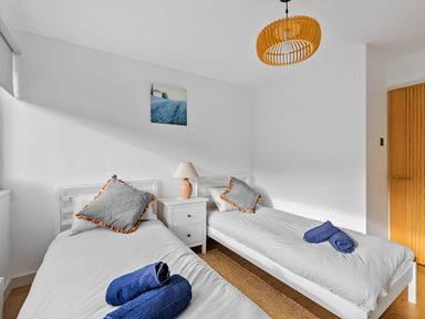 A bedroom with two single beds a small white nightstand with a lamp a wooden door and a painting on the wall at 5 Mermaid Court in Newquay
