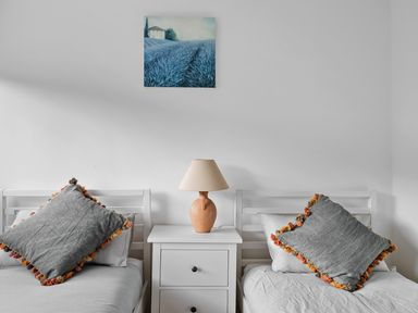 A bedroom with two single beds separated by a white nightstand with a lamp and a painting above at 5 Mermaid Court in Newquay