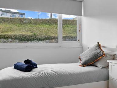 A single bed with a gray pillow and folded navy towels beside a window showing a grassy hill at 5 Mermaid Court in Newquay