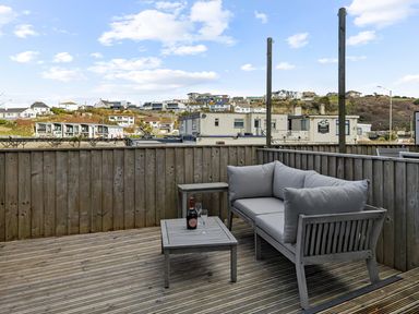 A wooden deck with a gray cushioned sofa and table with a bottle and glasses with buildings in the background at 5 Mermaid Court in Newquay