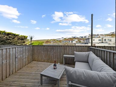A wooden balcony with a gray cushioned corner sofa and a small table with a bottle and two glasses at 5 Mermaid Court in Newquay