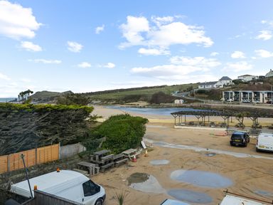 A sandy parking area with vehicles and picnic tables near a river and houses on a hill at 5 Mermaid Court in Newquay
