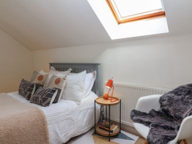 A bedroom with a bed multiple pillows a small side table with an orange lamp and a white chair with a fur throw at Priory Barn Apartment in Walton near Street