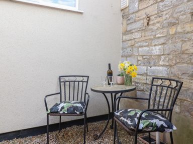 An outdoor seating area with two metal chairs with cushions a round table with a bottle of wine glasses and a vase of flowers at Priory Barn Apartment in Walton near Street