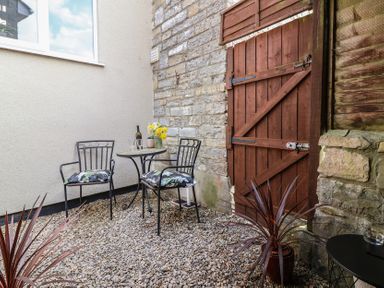 A small outdoor seating area with two metal chairs a round table with flowers and a bottle on gravel outside a stone wall and wooden door at Priory Barn Apartment in Walton near Street