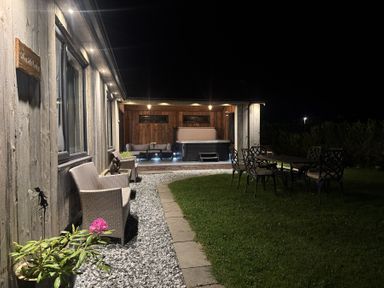 An outdoor patio at night with chairs on a gravel path a table with chairs on grass and a covered seating area with a hot tub at Wayside Lodge in Blackpool