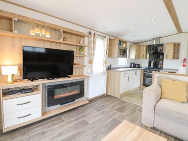 An open living area with a television above a fireplace lamp on a cabinet kitchen with cabinets and appliances and a sofa with a yellow cushion at 75 Foxburrow Hang in Fritton