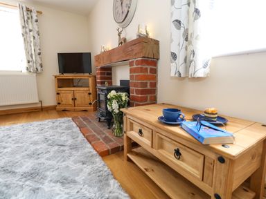 A living room with a wooden cabinet tv a fireplace with flowers on the hearth and a wooden coffee table with a book a cup and a plate near a window with patterned curtains at Little Dairy House near Swanage