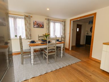 A dining area with a wooden table and four chairs on a rug near two windows with curtains at Little Dairy House near Swanage